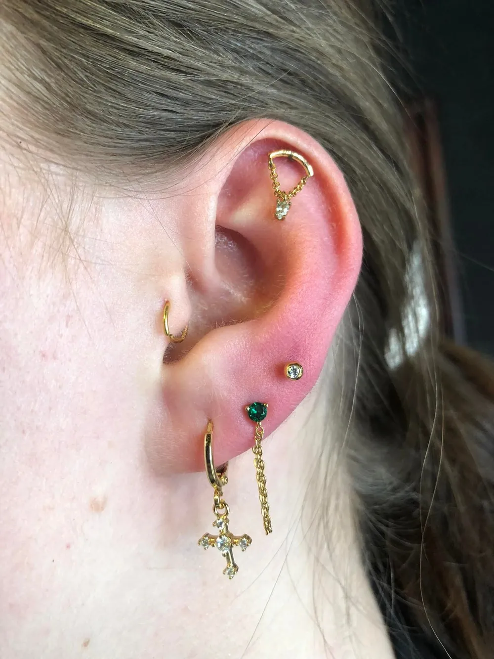 Oor met een gouden flat juweel met kettinkje, een tragus ring en meerdere oorlelpiercings met een groene steen en een kruisje